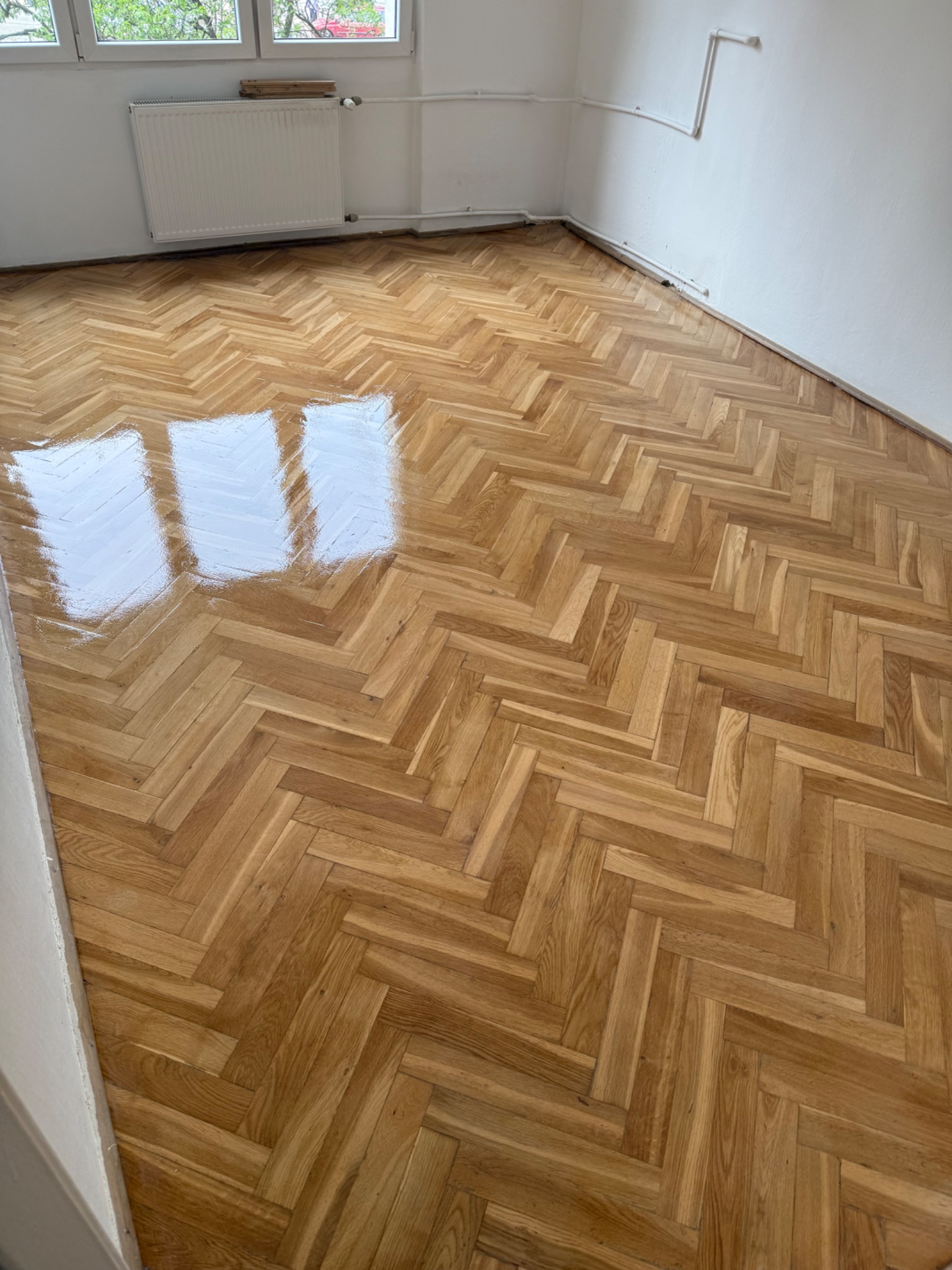 Świeżo polakierowany parkiet ułożony w jodełkę, odbijający światło z okna, w pustym, odnowionym pokoju z białymi ścianami i grzejnikiem.