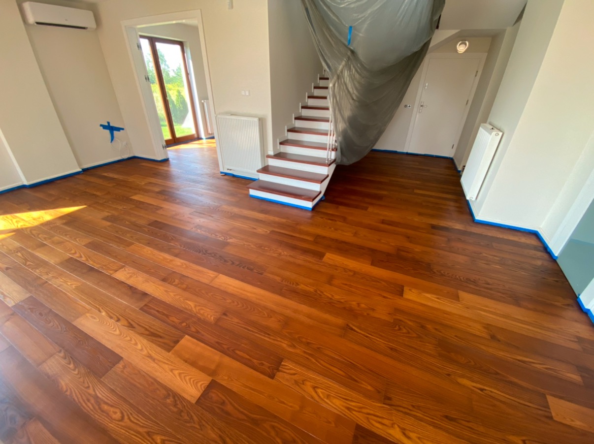 Wycyklinowany, lakierowany parkiet w salonie z zabezpieczonymi taśmą ścianami i schodami pod folią ochronną. Widoczne naturalne usłojenie drewna.