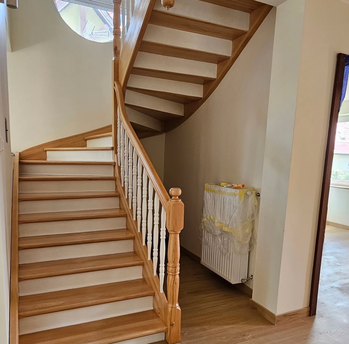 Drewniane schody spiralne z białymi podstopnicami, widoczne z dołu, z okrągłym oknem w tle. Jasne drewno, białe balustrady i radiator z osłoną.