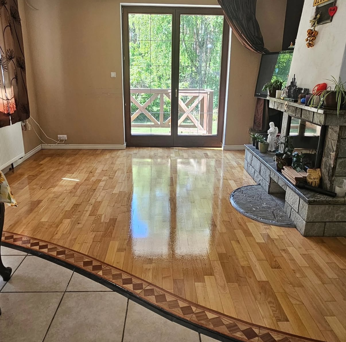 Błyszcząca, odnowiona podłoga parkietowa w salonie z kominkiem i wyjściem na balkon, wykończona ozdobną bordiurą. Widoczne refleksy światła na lakierowanej powierzchni.