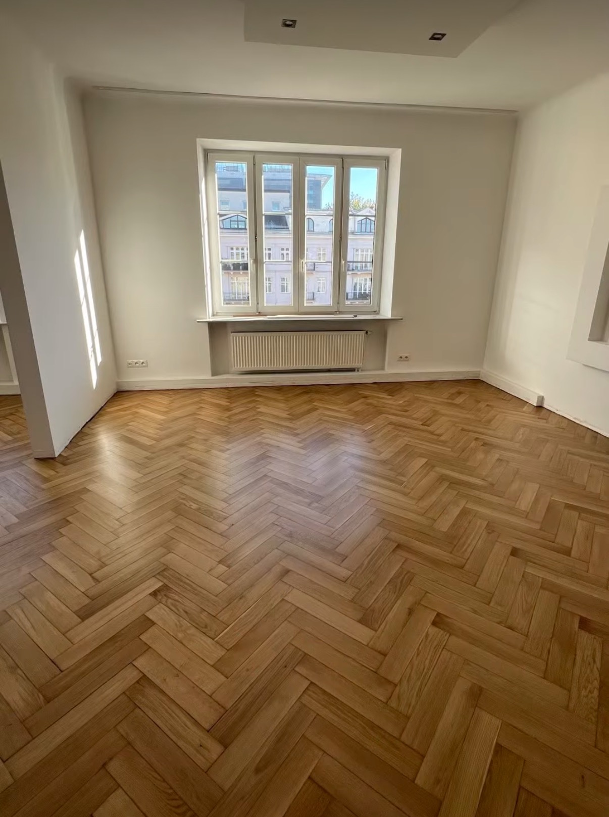 Jasny parkiet ułożony w jodełkę w przestronnym pokoju z oknem. Widoczny grzejnik pod oknem i fragment kolumny. Naturalne światło podkreśla strukturę drewna.