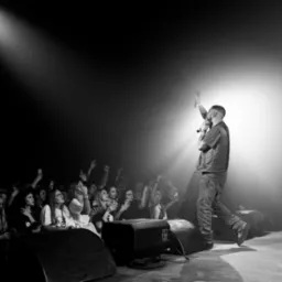 Czarno-białe zdjęcie z koncertu. Wokalista na scenie, oświetlony reflektorem, a przed nim rozentuzjazmowany tłum fanów z podniesionymi rękami i telefonami.