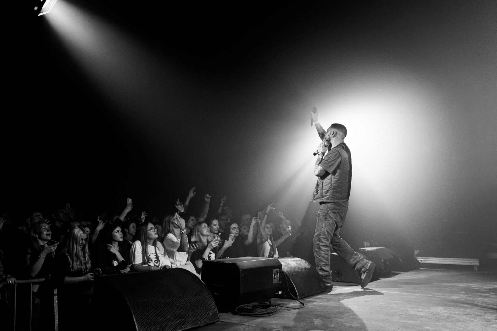 Czarno-białe zdjęcie z koncertu. Wokalista na scenie, oświetlony reflektorem, a przed nim rozentuzjazmowany tłum fanów z podniesionymi rękami i telefonami.
