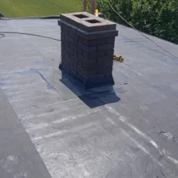 Murarz w czerwonej koszulce i ciemnych spodenkach pracuje na płaskim dachu, budując komin z cegły. W tle widać zielone pole i kominy fabryczne.