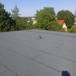 VOVABUD - Szary, płaski dach pokryty papą termozgrzewalną, z widocznym kominkiem wentylacyjnym i otoczeniem zieleni oraz zabudowaniami w tle, sfotografowany w słoneczny dzień.