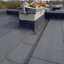 VOVABUD - Widok na płaski dach pokryty papą, z kominem wentylacyjnym i stertą materiałów budowlanych na jego szczycie, w tle drzewa bez liści.