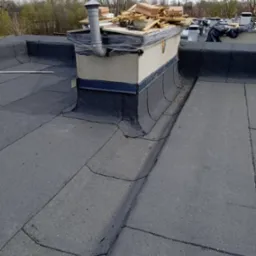 Widok na płaski dach pokryty papą, z kominem wentylacyjnym i stertą materiałów budowlanych na jego szczycie, w tle drzewa bez liści.