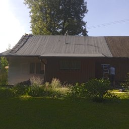 VOVABUD - Prace na dachu domu jednorodzinnego w trakcie realizacji, widoczny fragment dachu pokryty papą, drabina oparta o dach, pracownik na drabinie, bujna roślinność w ogrodzie.