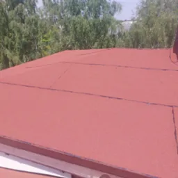 Czerwony dach pokryty papą na tle zieleni drzew i błękitnego nieba. Widoczne rynny. Nowe pokrycie dachowe z bliska w słoneczny dzień.