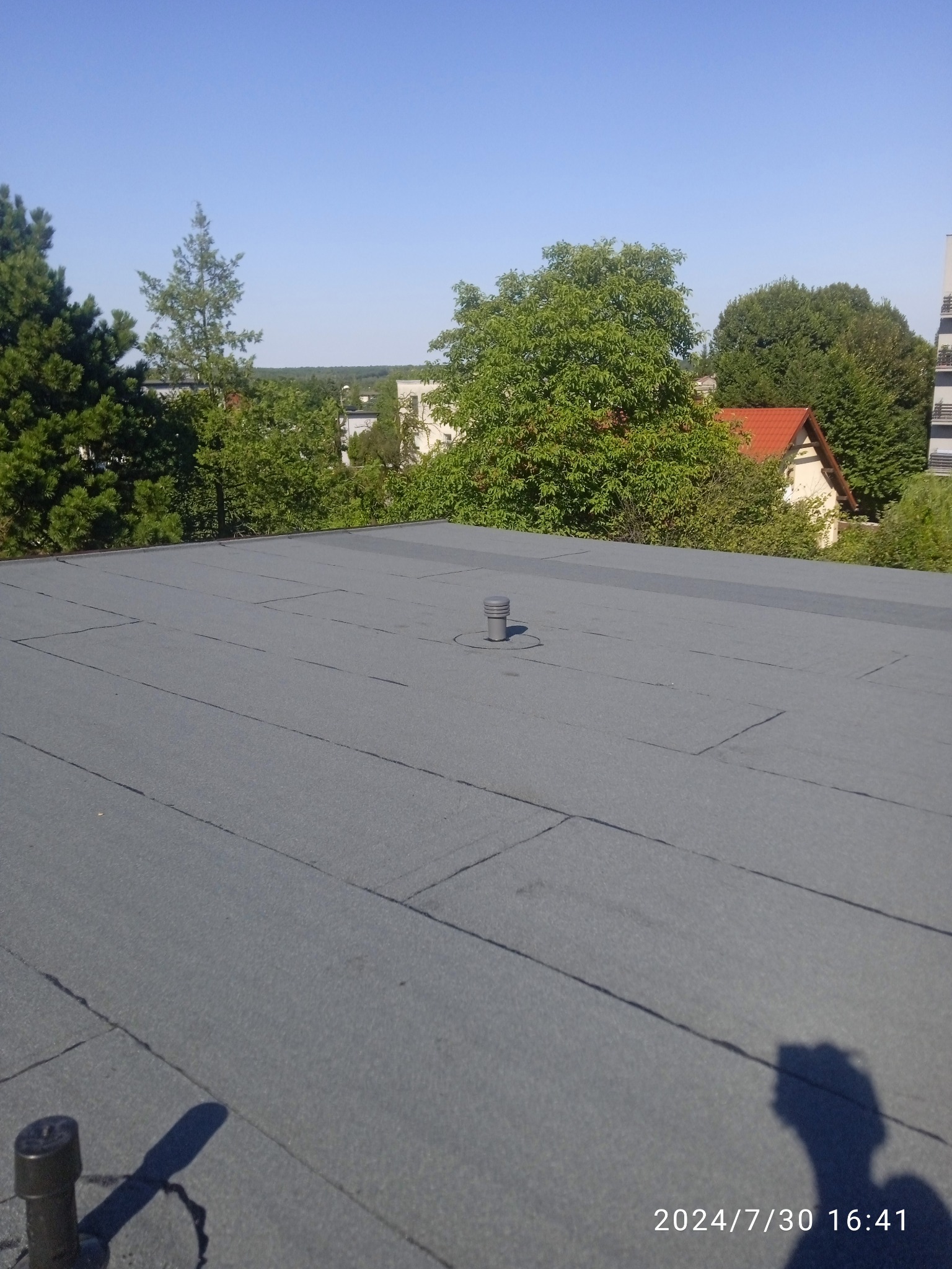 Szary, płaski dach pokryty papą termozgrzewalną, z widocznym kominkiem wentylacyjnym i otoczeniem zieleni oraz zabudowaniami w tle, sfotografowany w słoneczny dzień.