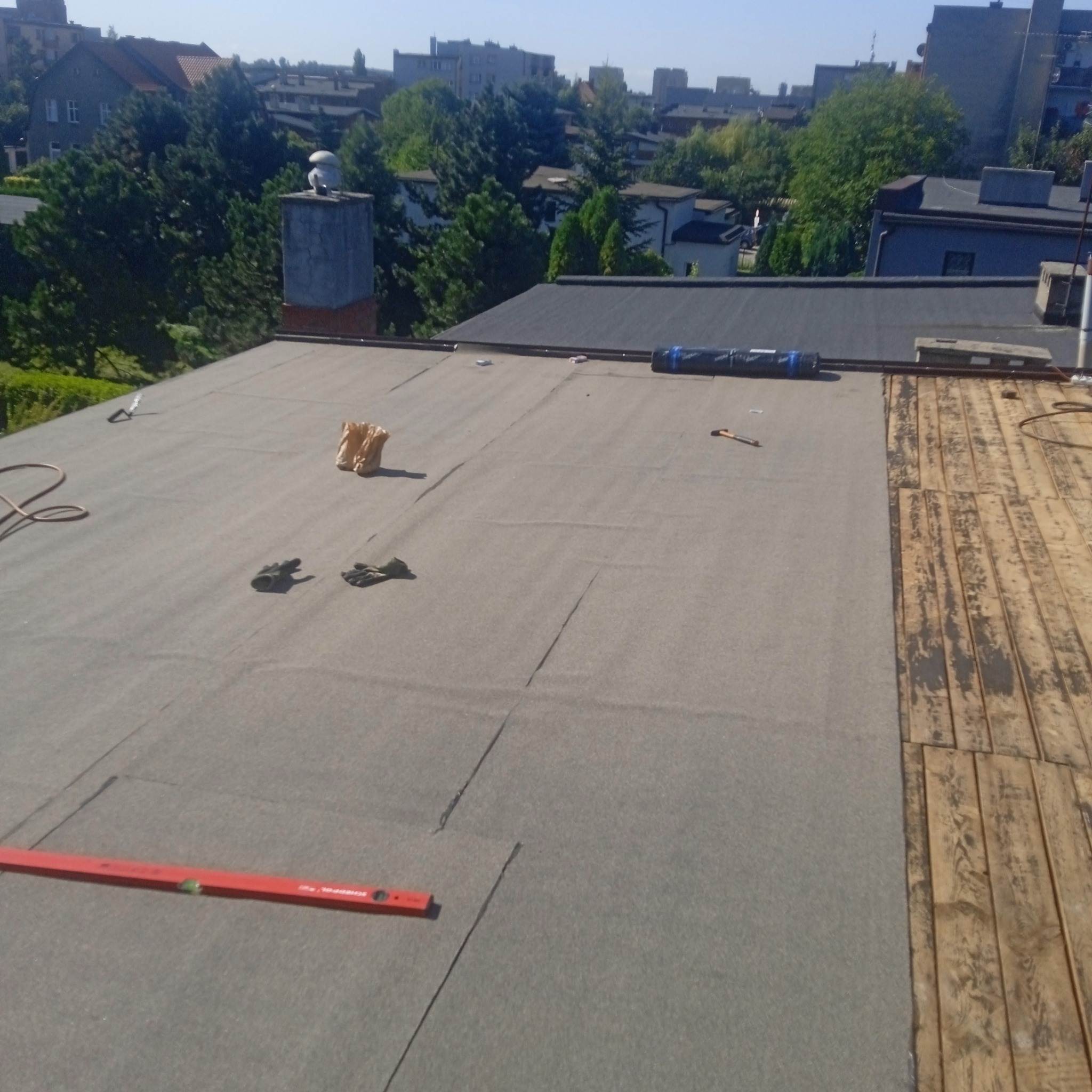 Fragment dachu z papy termozgrzewalnej, częściowo pokrytego nową warstwą, obok odsłonięte deski poszycia, poziomica, rękawice robocze i rolka papy w tle, widok z góry na tle zabudowy miejskiej...