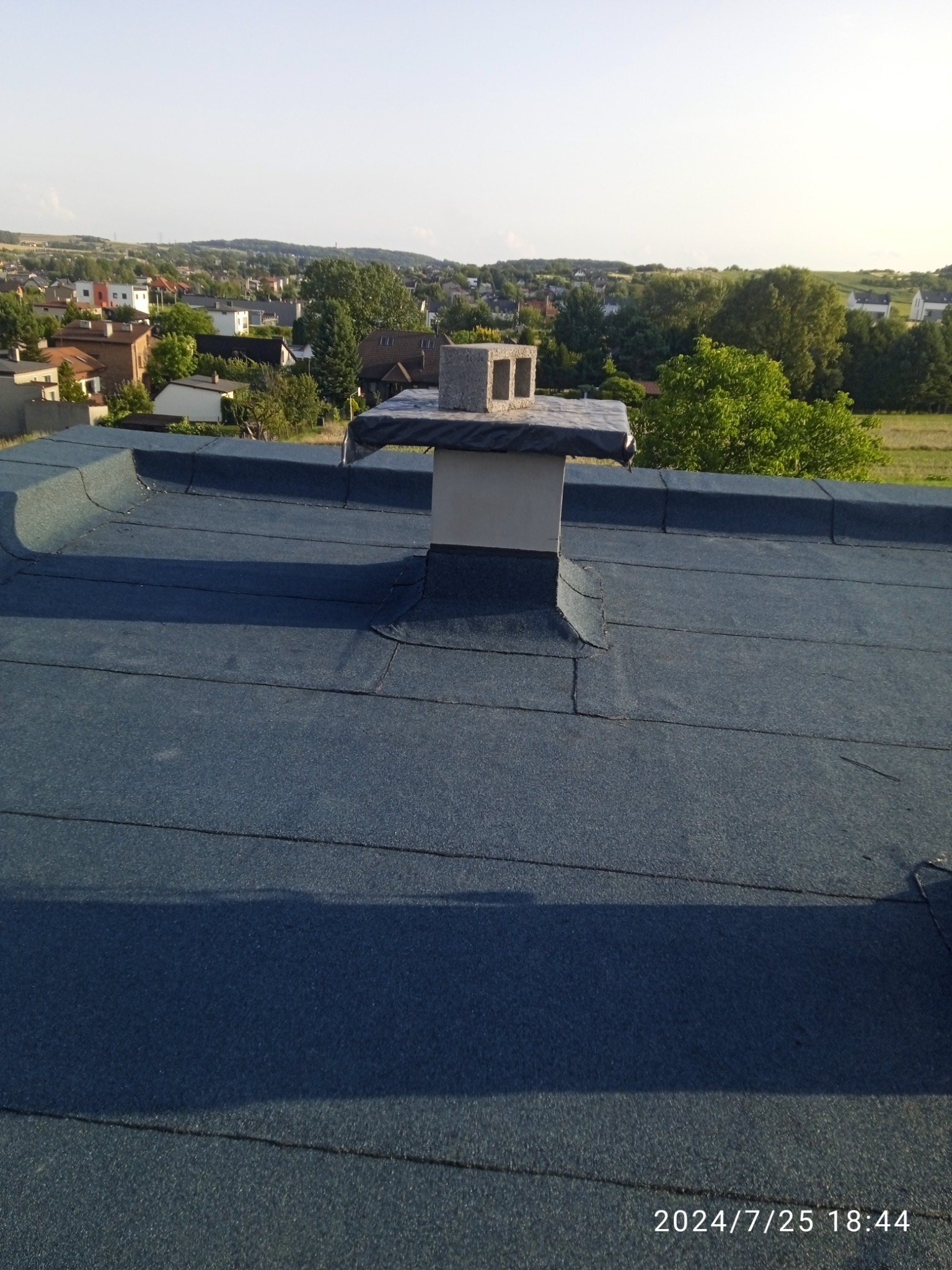 Niebieski dach pokryty papą z kominem wentylacyjnym zakończonym betonowymi pustakami, w tle zabudowa jednorodzinna i zieleń.