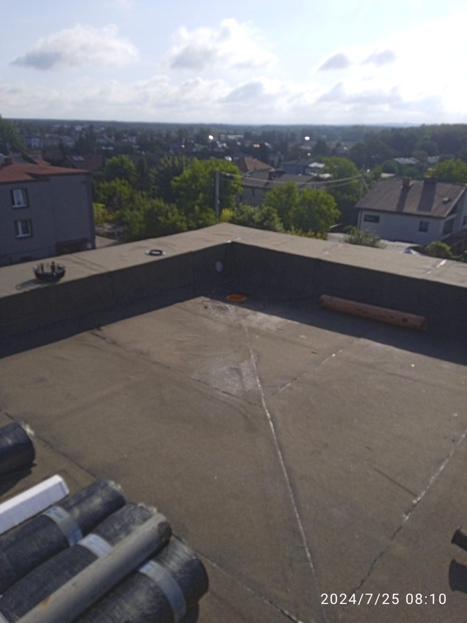 Widok z góry na płaski dach pokryty papą, z widocznymi rolkami materiału izolacyjnego, elementami wentylacyjnymi i panoramą miasta w tle.