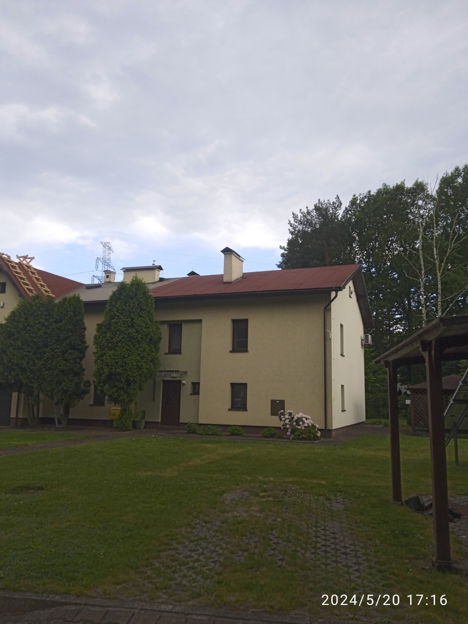 Dwupiętrowy budynek z czerwonym dachem, częściowo zdjętym, z widoczną konstrukcją drewnianą i drabiną, otoczony zielonymi drzewami i trawnikiem w pochmurny dzień.