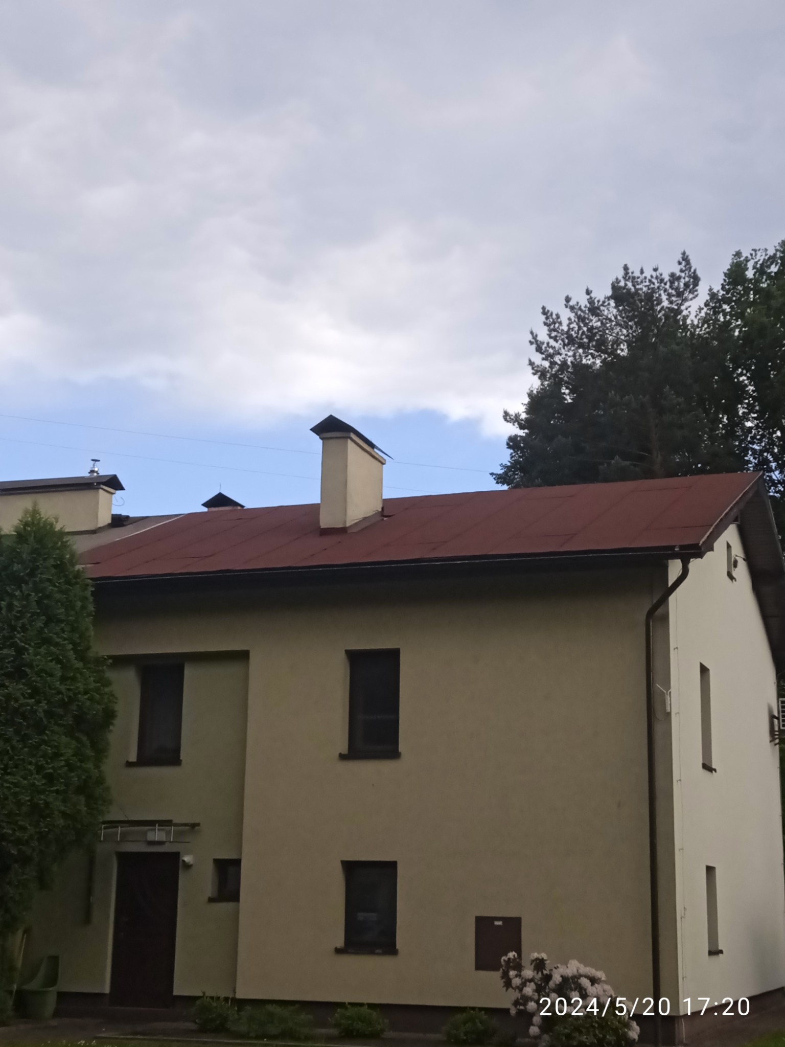 Dwupiętrowy dom z czerwonym dachem z blachy, widoczny z poziomu gruntu w pochmurny dzień. Na dachu widoczne kominy. Budynek otynkowany, w kolorze beżowym i białym. Widoczne rynny.