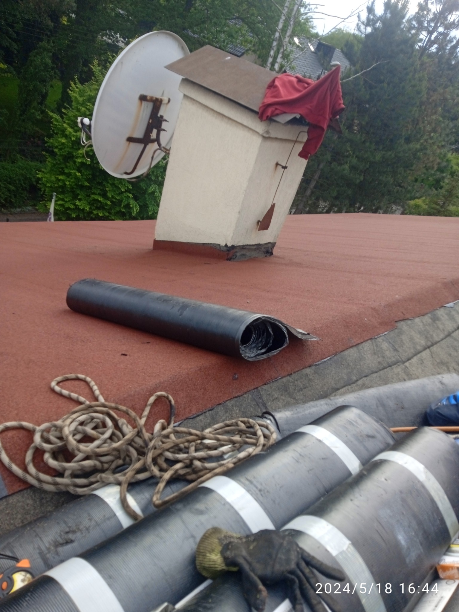 Czerwony dach pokryty papą z kominem, anteną satelitarną, zwiniętym kawałkiem papy, rolkami papy, liną i roboczą rękawicą.