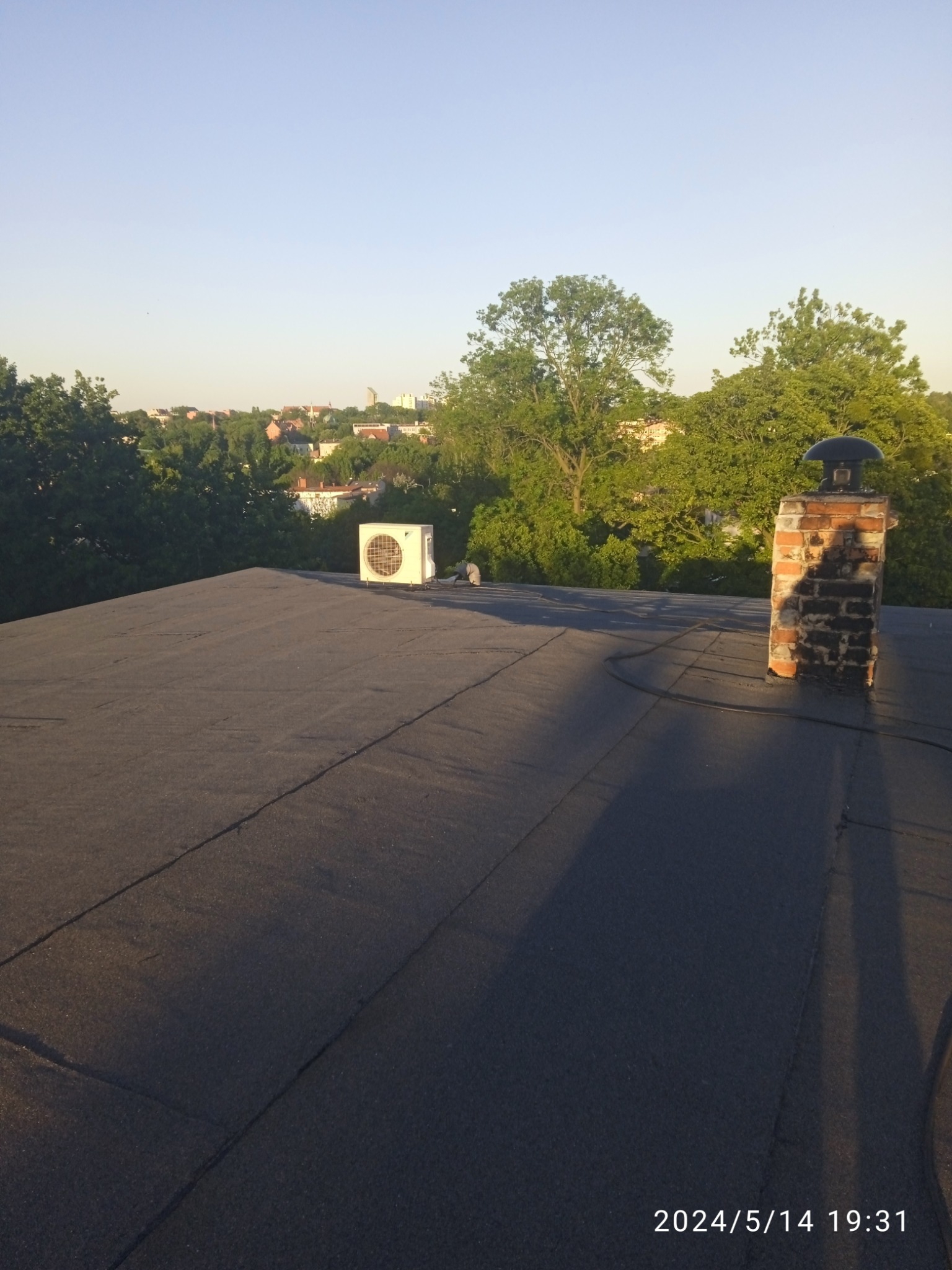 Widok na płaski dach pokryty papą z widocznym kominem z cegły, klimatyzatorem i panoramą miasta w tle, sfotografowany o zachodzie słońca.