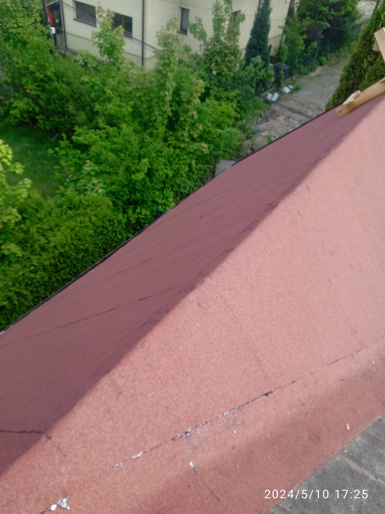Czerwony dach pokryty papą bitumiczną, widok z góry, z fragmentem łat drewnianych w prawym górnym rogu, na tle zieleni ogrodowej i zabudowań.