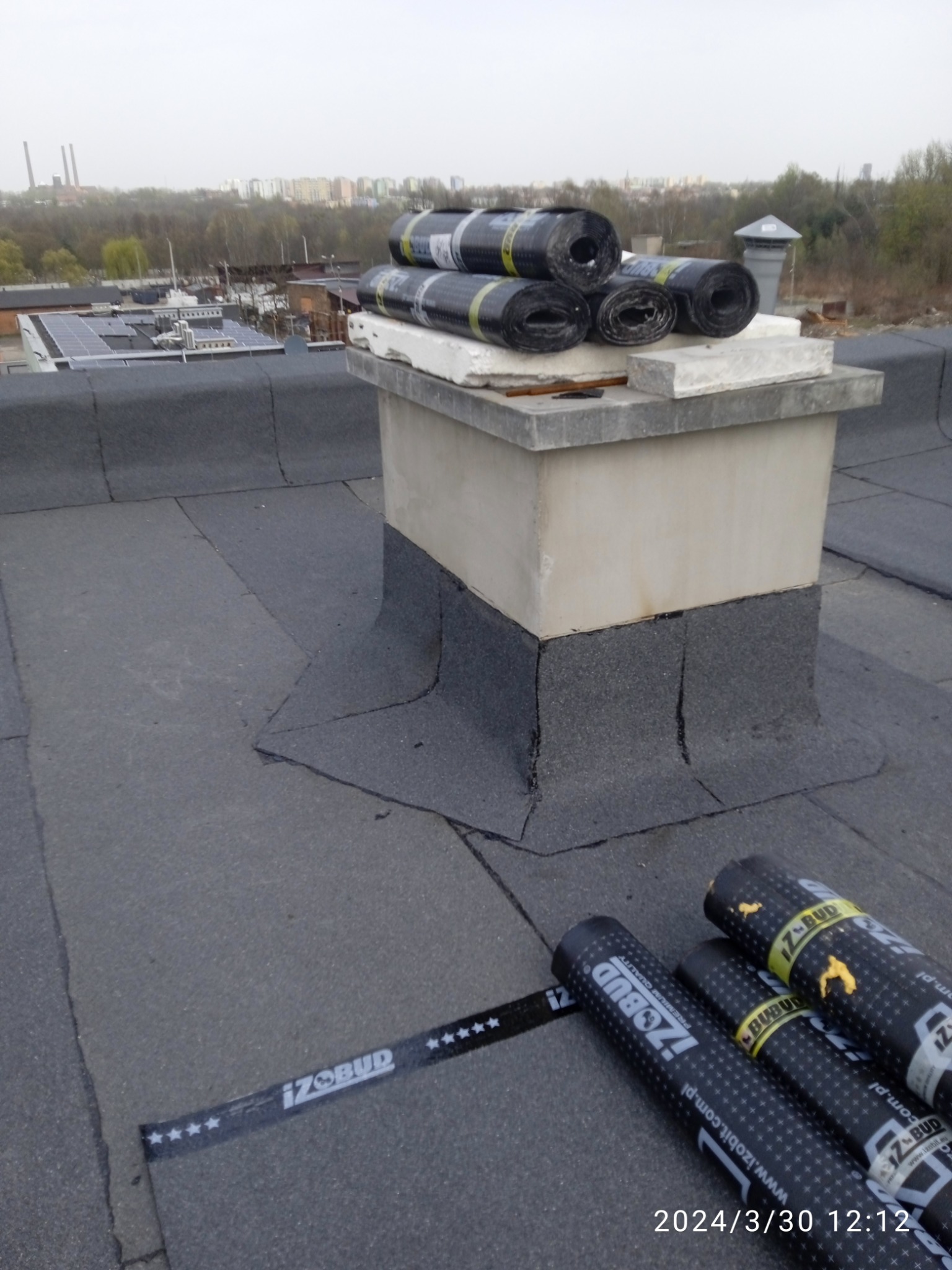 Widok z góry na płaski dach pokryty papą, z kominem obłożonym papą i kilkoma rolkami papy Izobud na wierzchu oraz na dachu, z widokiem na zabudowania przemysłowe i drzewa w tle.