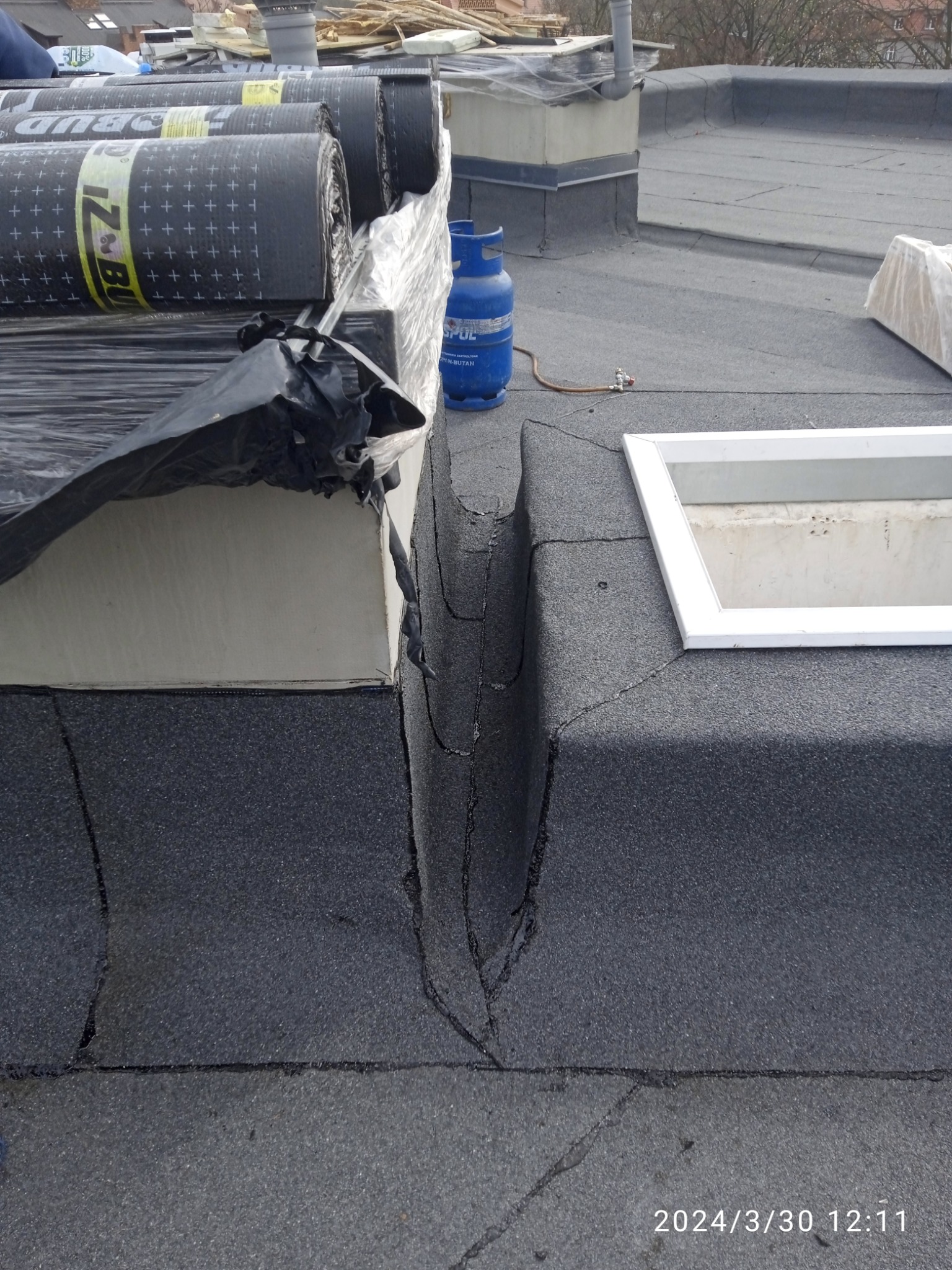 Płaski dach pokryty papą, z widocznym kominem, świetlikiem i szczeliną dylatacyjną wypełnioną masą bitumiczną, obok przygotowane rolki papy i butla gazowa.