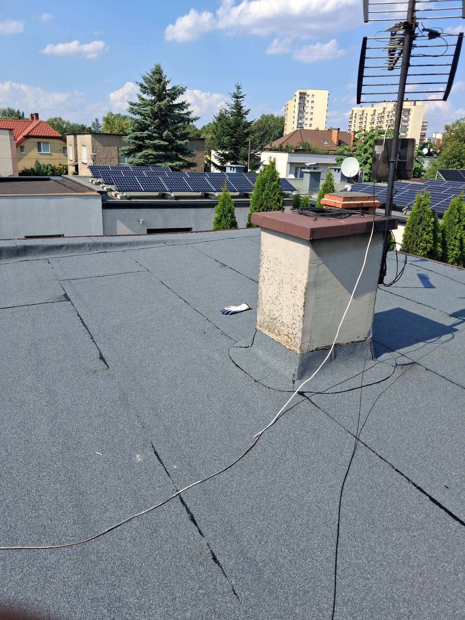 Płaski dach pokryty papą z widocznym kominem, przewodami i pojedynczą rękawicą roboczą, w tle zabudowa miejska i panele słoneczne.