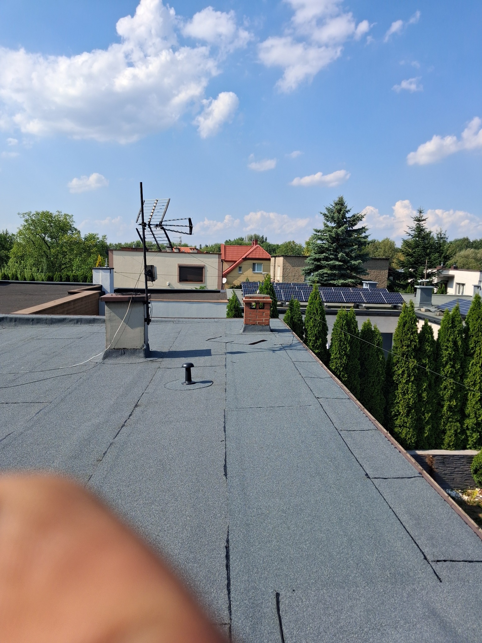 Szary, płaski dach pokryty papą termozgrzewalną z widocznym kominem, anteną telewizyjną i panelami słonecznymi na sąsiednich budynkach w tle.