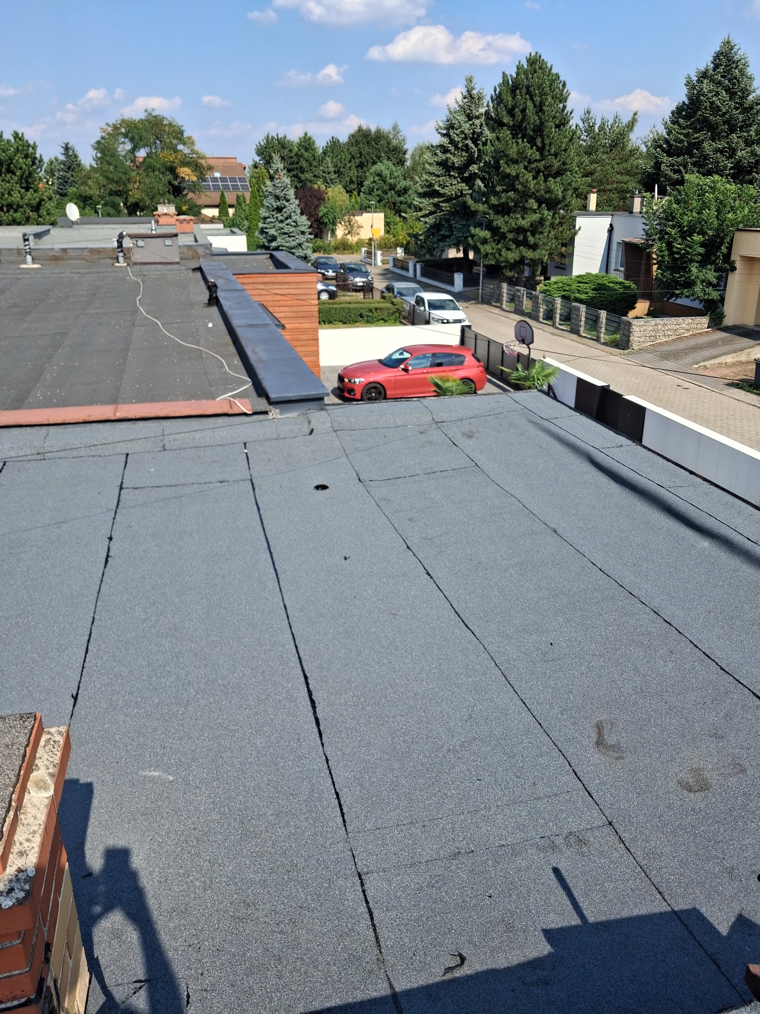 Widok z góry na płaski dach pokryty papą, z widocznym czerwonym samochodem osobowym zaparkowanym obok budynku na tle zieleni i błękitnego nieba.