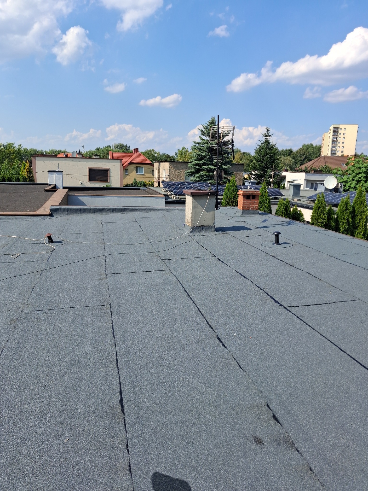 Płaski dach pokryty papą termozgrzewalną z widocznymi kominami, odpowietrznikiem i anteną na tle zabudowy jednorodzinnej w słoneczny dzień.