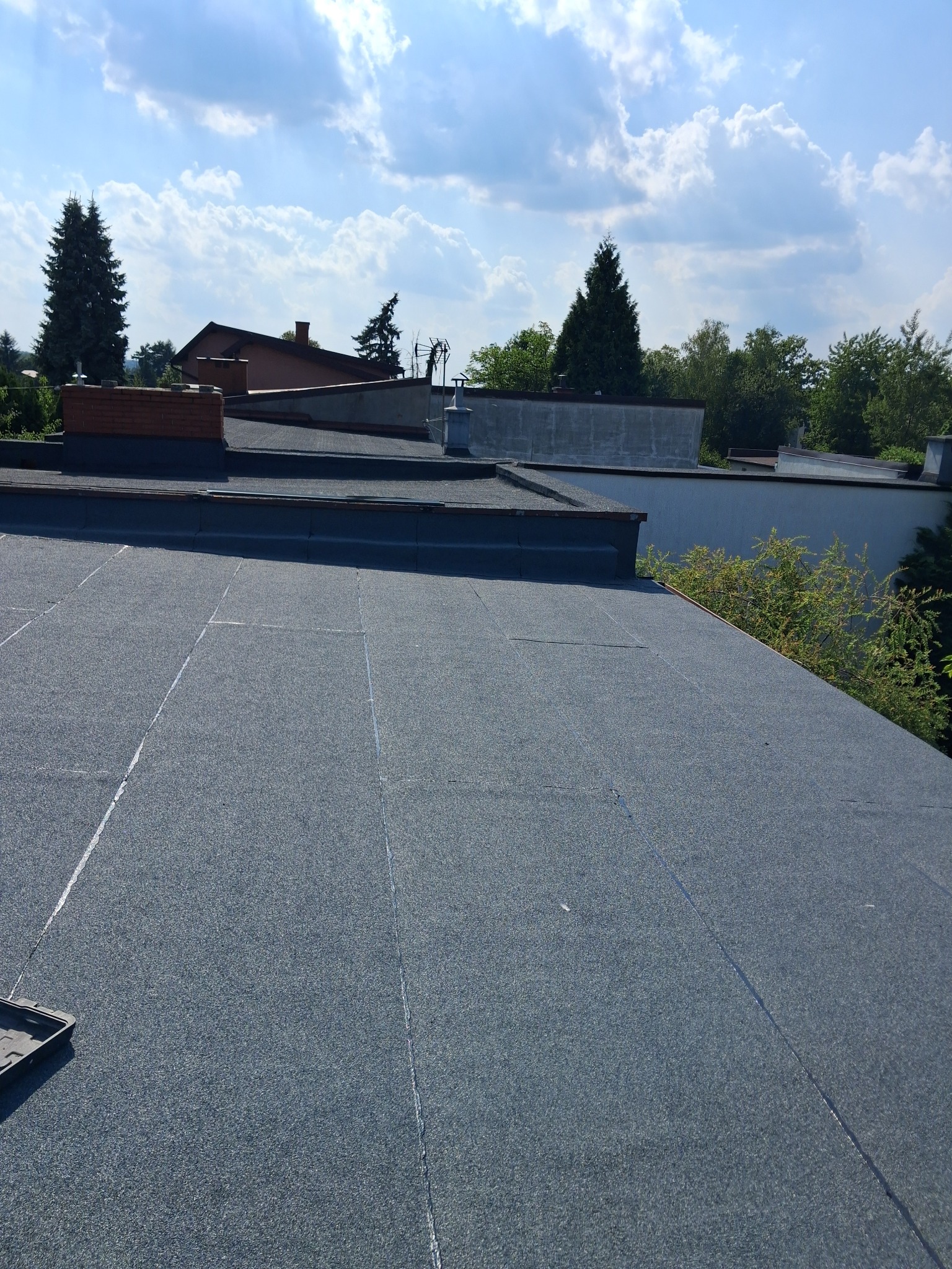 Widok z góry na płaski dach pokryty papą z widocznymi łączeniami, w tle budynki i drzewa pod błękitnym niebem z chmurami.