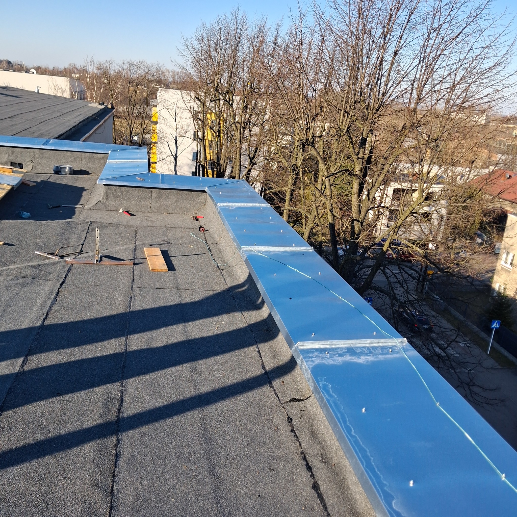 Nowe, błyszczące, metalowe obróbki blacharskie na płaskim dachu pokrytym papą, widoczne narzędzia i drewniane elementy, w tle drzewa i zabudowa miejska.