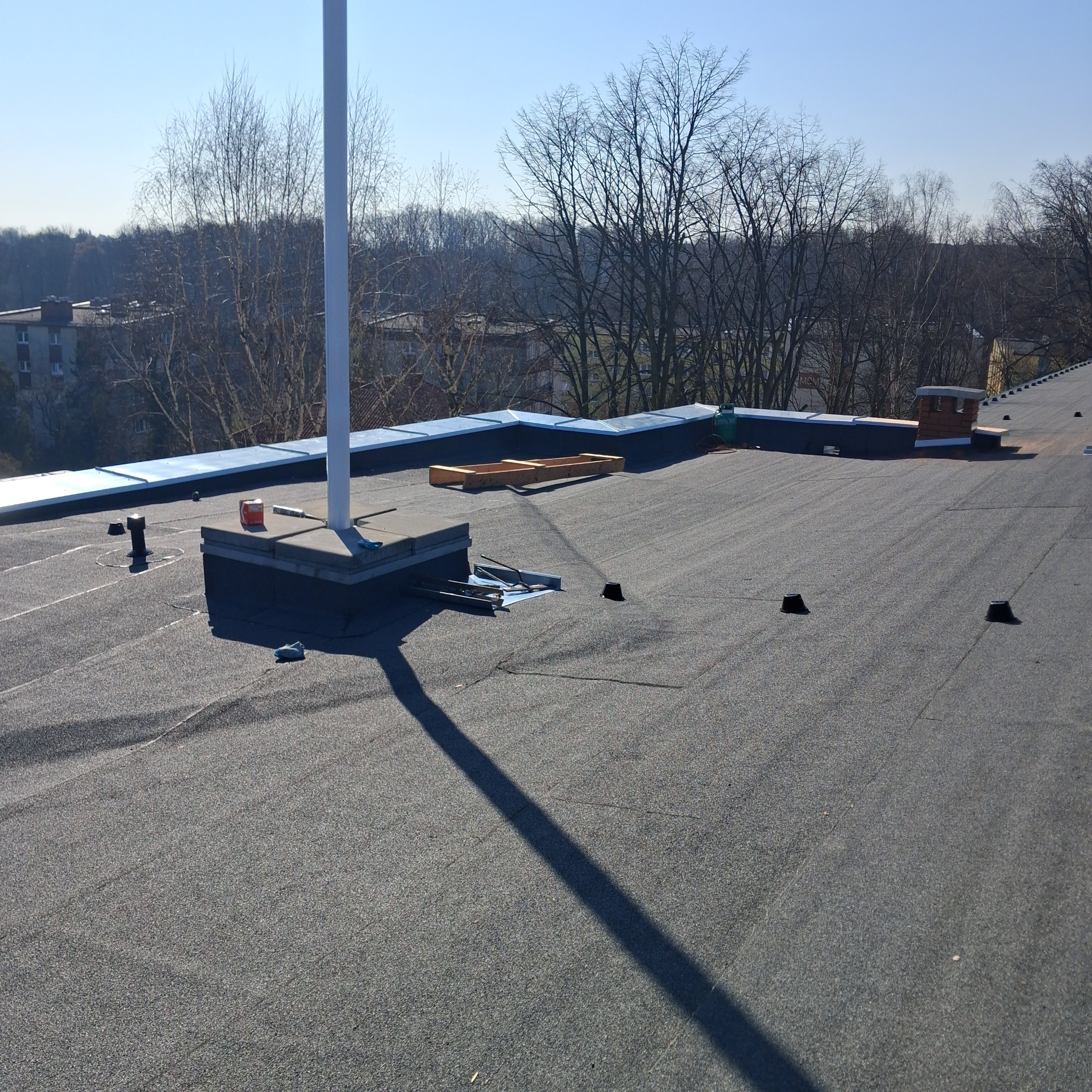 Płaski dach pokryty papą termozgrzewalną z widocznymi elementami wentylacyjnymi, kominem i narzędziami, w tle drzewa i budynek mieszkalny.