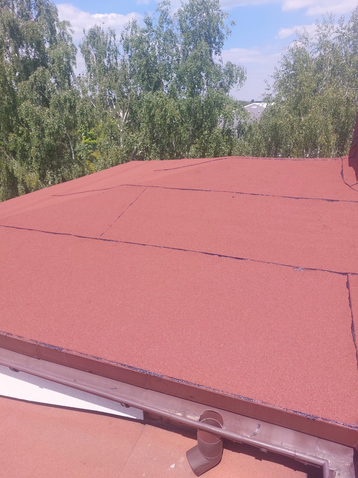 Czerwony dach pokryty papą na tle zieleni drzew i błękitnego nieba. Widoczne rynny. Nowe pokrycie dachowe z bliska w słoneczny dzień.
