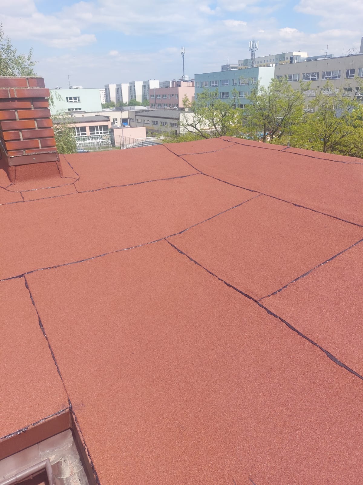 Czerwony dach pokryty papą termozgrzewalną z widocznym kominem i fragmentem rynny na tle budynków miejskich w słoneczny dzień.