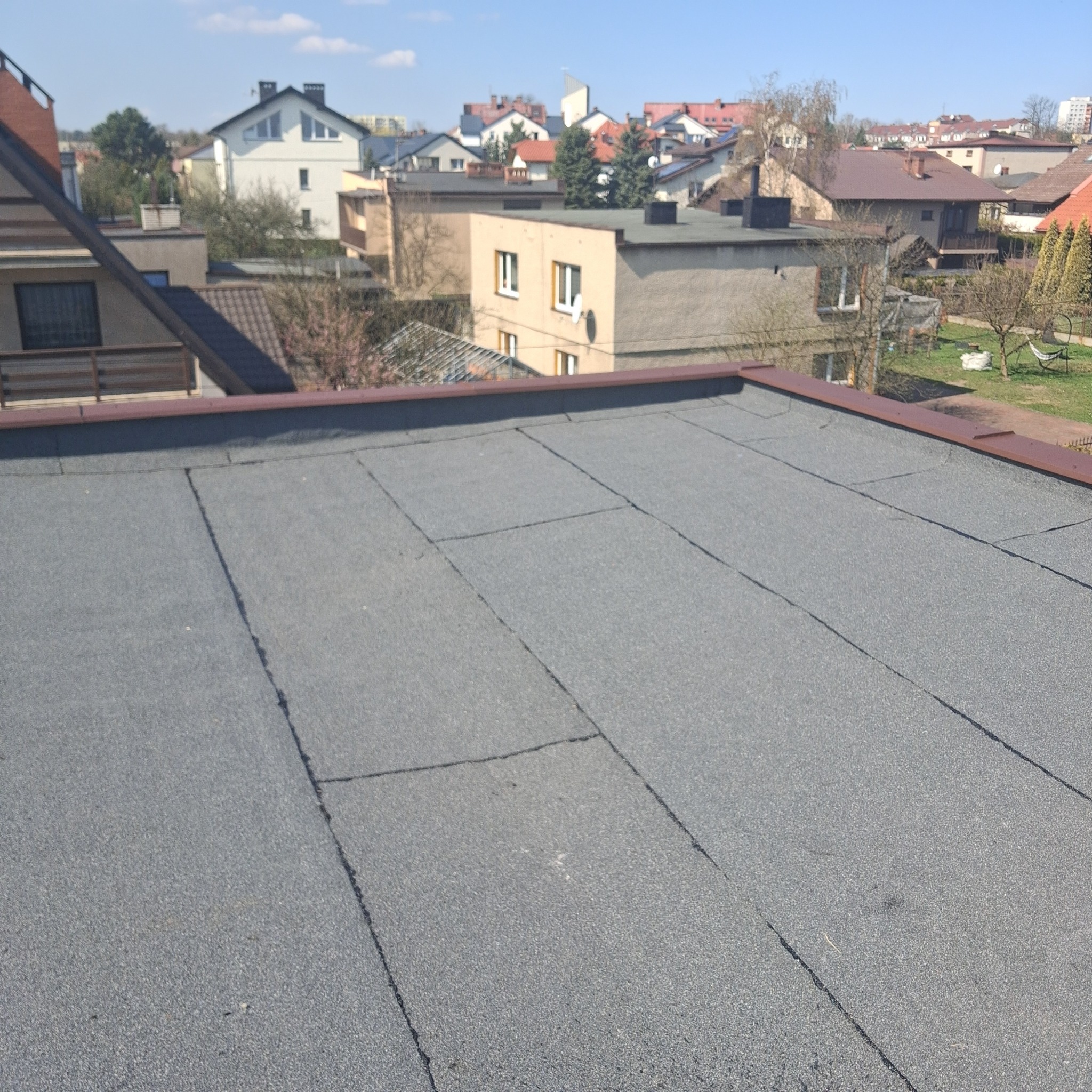 Szary, płaski dach pokryty papą na tle zabudowy miejskiej w słoneczny dzień. Widoczne łączenia papy i brązowe obrzeże dachu. Perspektywa z góry.
