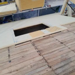 Montaż izolacji termicznej na dachu z wyciętym otworem, widoczne deski starego dachu i przygotowane płyty izolacyjne na rusztowaniu.