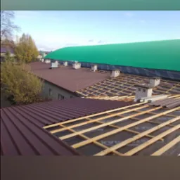 Widok z góry na konstrukcję dachu w trakcie budowy, z widocznymi łatami drewnianymi na czarnej membranie, fragmenty pokrycia z bordowej blachy trapezowej oraz kominy wentylacyjne. W tle budynek...