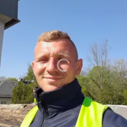 Uśmiechnięty dekarz w odblaskowej kamizelce robi selfie na dachu, w tle fragment budynku i drzewa.