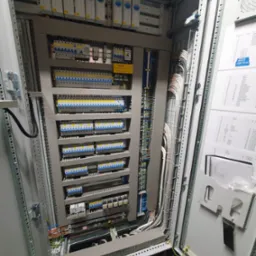 Otwarta szafa sterownicza z widocznymi bezpiecznikami, modułami, kablami i dokumentacją techniczną wewnątrz, z żółtą naklejką ostrzegawczą na górze i wentylatorem po prawej stronie.