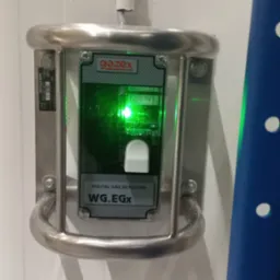 Zamontowany detektor gazu GAZEX WG.EGx w metalowej obudowie ochronnej, z widoczną świecącą diodą sygnalizacyjną, zainstalowany na białej ścianie chłodni.