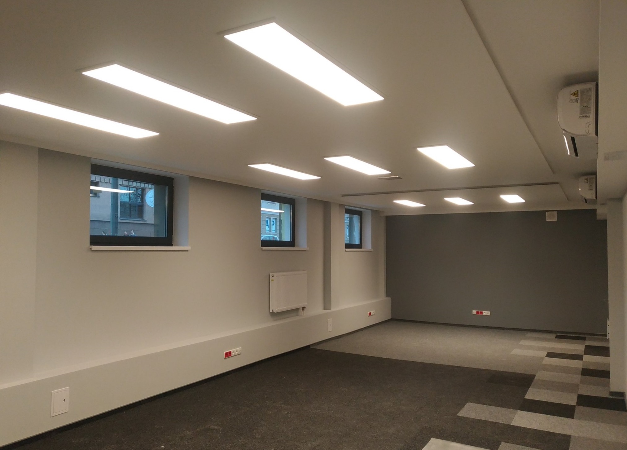 Wnętrze biurowe po remoncie z zainstalowanym oświetleniem LED i klimatyzacją, widoczne okna, grzejnik i gniazdka elektryczne w listwach przypodłogowych.