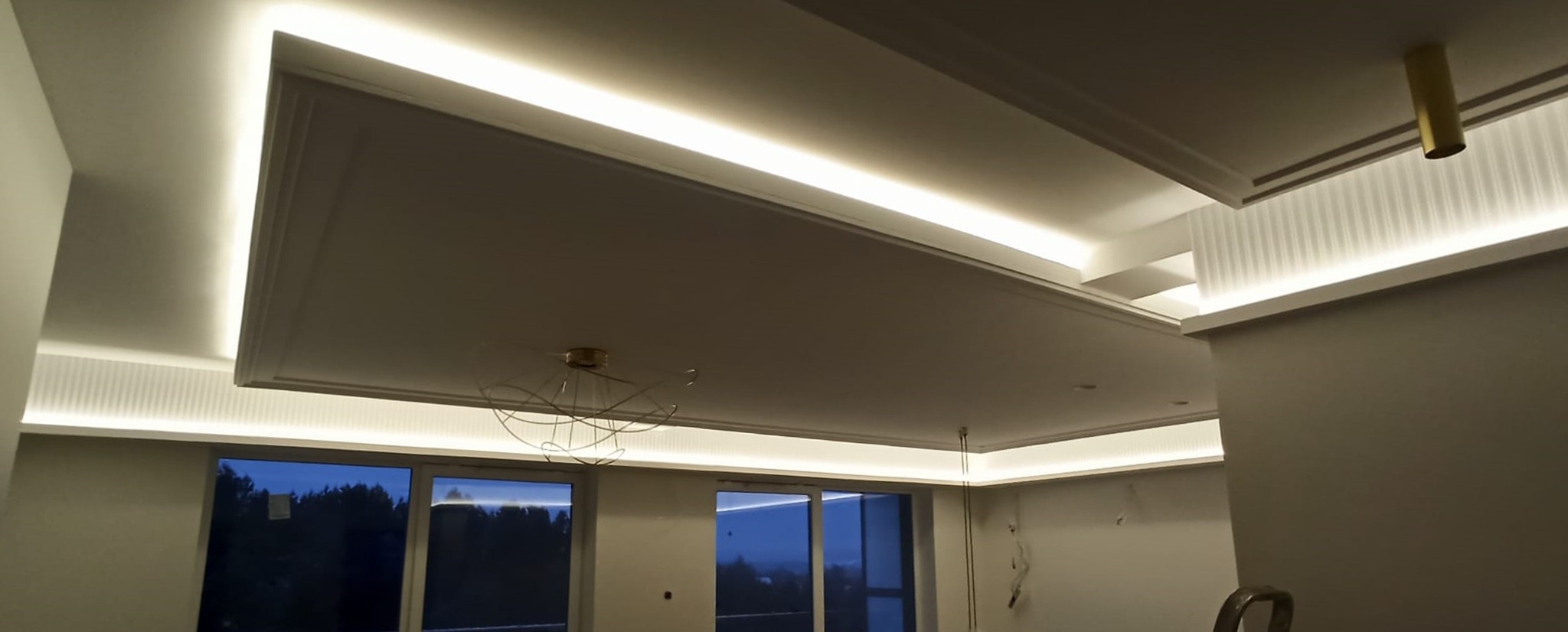 Nowoczesny sufit z podwieszanymi elementami, oświetleniem LED wzdłuż krawędzi i ozdobną lampą wiszącą, widok na okna z widokiem na drzewa w tle.