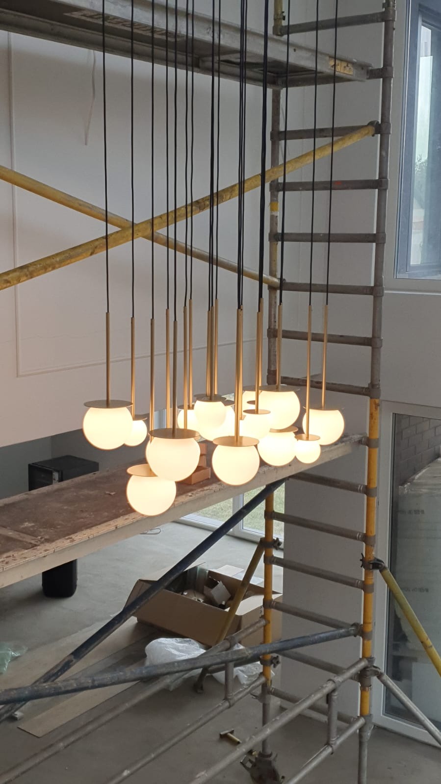 Montaż szklanych kulistych lamp wiszących z metalowymi elementami na czarnych kablach, widoczne rusztowanie malarskie.