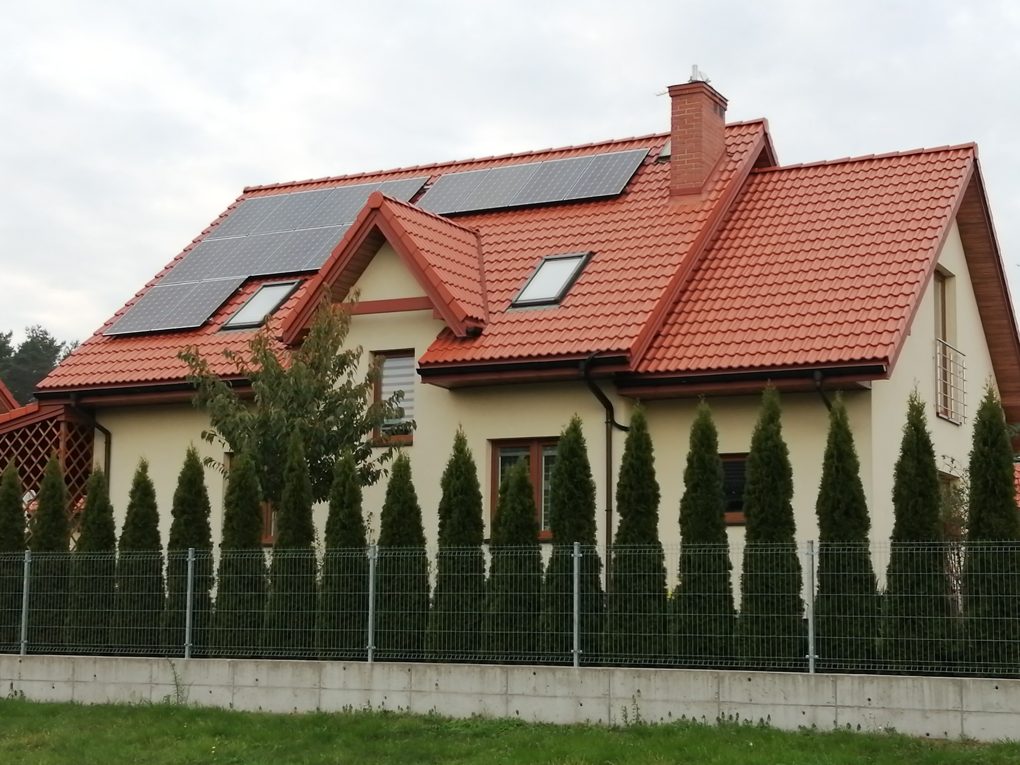 Dom jednorodzinny z panelami fotowoltaicznymi na dachu z czerwonej dachówki, widoczne okna dachowe i komin, zielone tuje wzdłuż metalowego ogrodzenia.