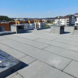 Szary, płaski dach pokryty papą termozgrzewalną na tle nowoczesnego osiedla w słoneczny dzień.