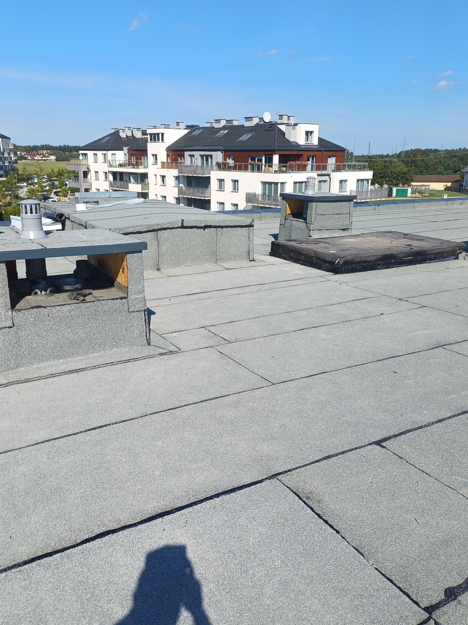 Szary, płaski dach z papy termozgrzewalnej na tle nowoczesnego budynku mieszkalnego w słoneczny dzień. Widoczne kominy wentylacyjne i cień osoby robiącej zdjęcie.