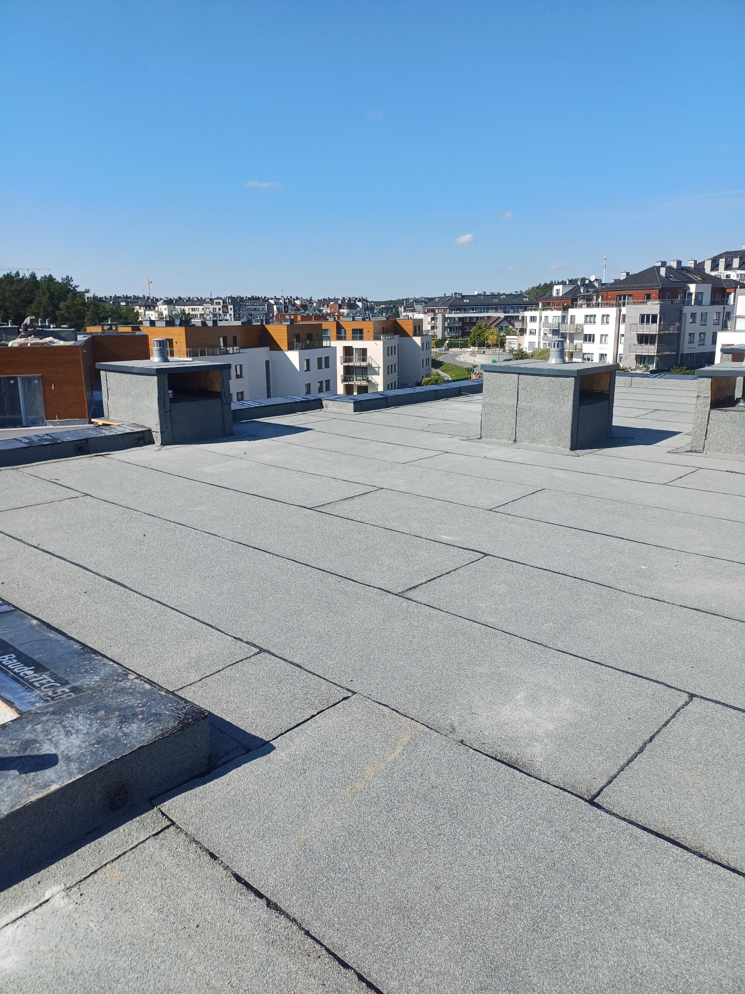Szary, płaski dach pokryty papą termozgrzewalną na tle nowoczesnego osiedla w słoneczny dzień.