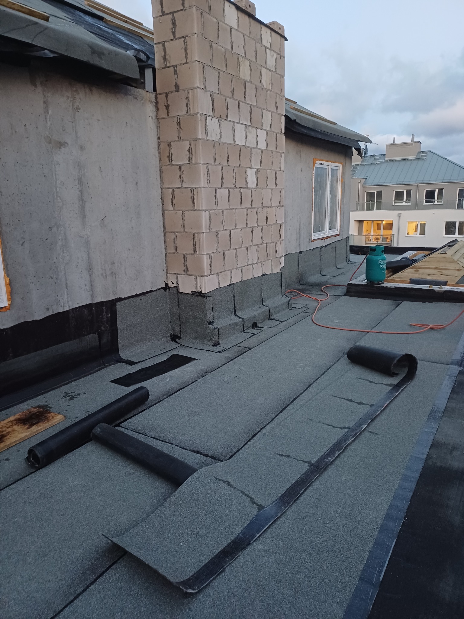 Widok z góry na płaski dach pokryty papą termozgrzewalną, częściowo zakończony, z widocznym kominem z bloczków betonowych, butlą gazową i narzędziami, na tle budynku mieszkalnego.