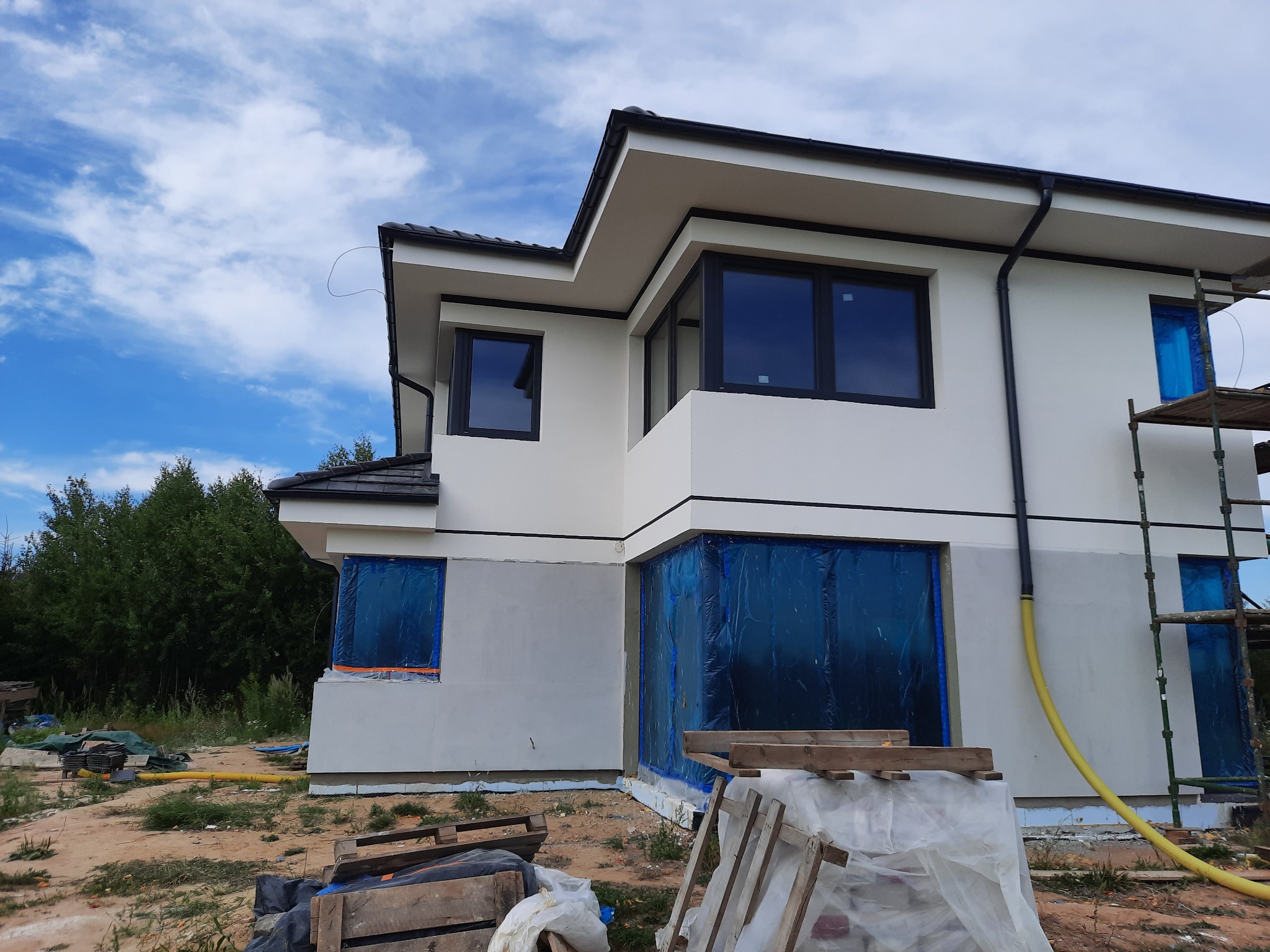 Nowoczesny dom w trakcie prac elewacyjnych: część ściany pomalowana na biało, okna zabezpieczone folią, widoczne rusztowania i węże, teren wokół w trakcie porządkowania.