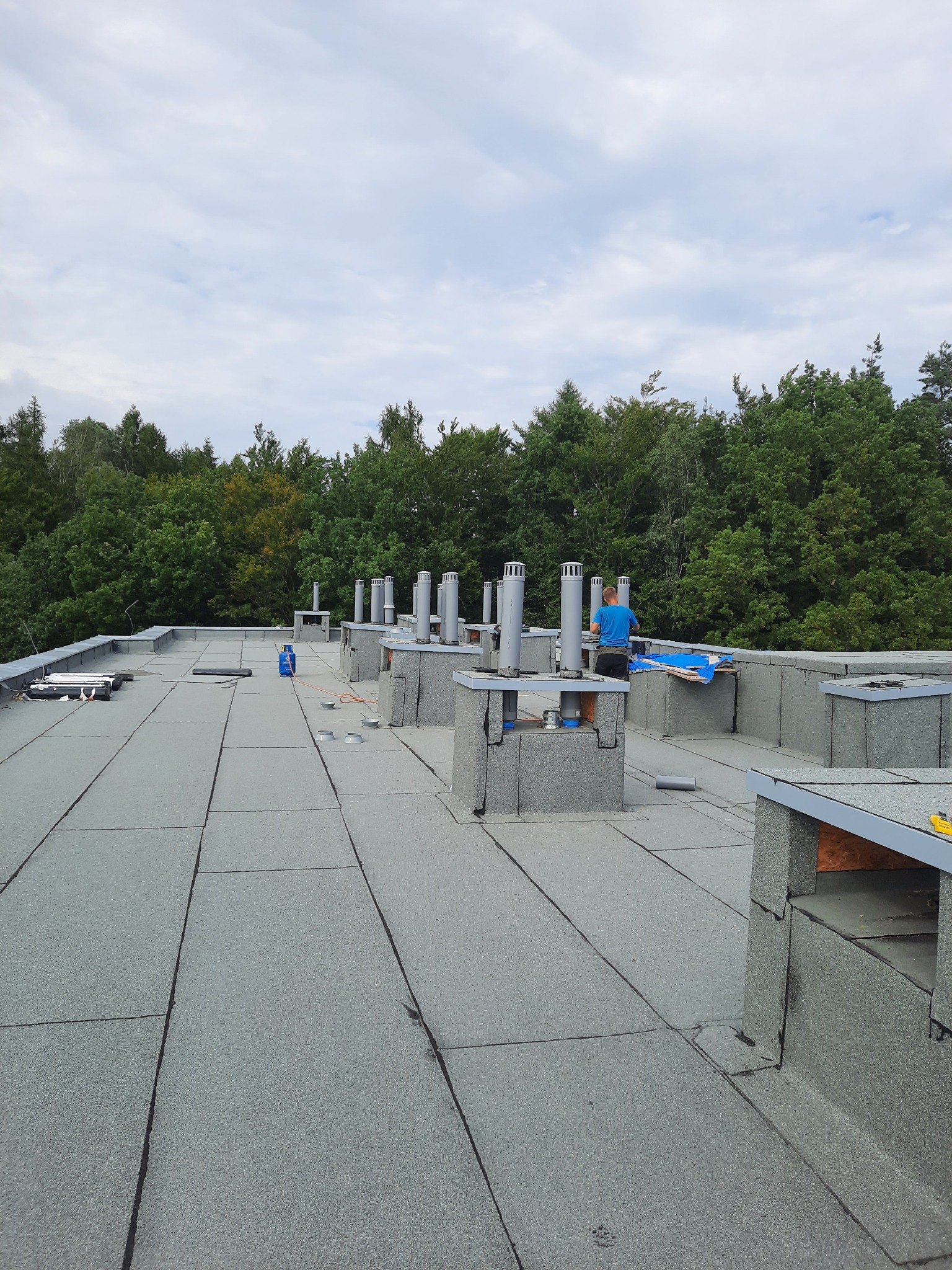 Wykończony płaski dach pokryty papą termozgrzewalną z obróbkami blacharskimi kominów wentylacyjnych, w tle drzewa i pochmurne niebo. Widoczny pracownik w trakcie montażu.