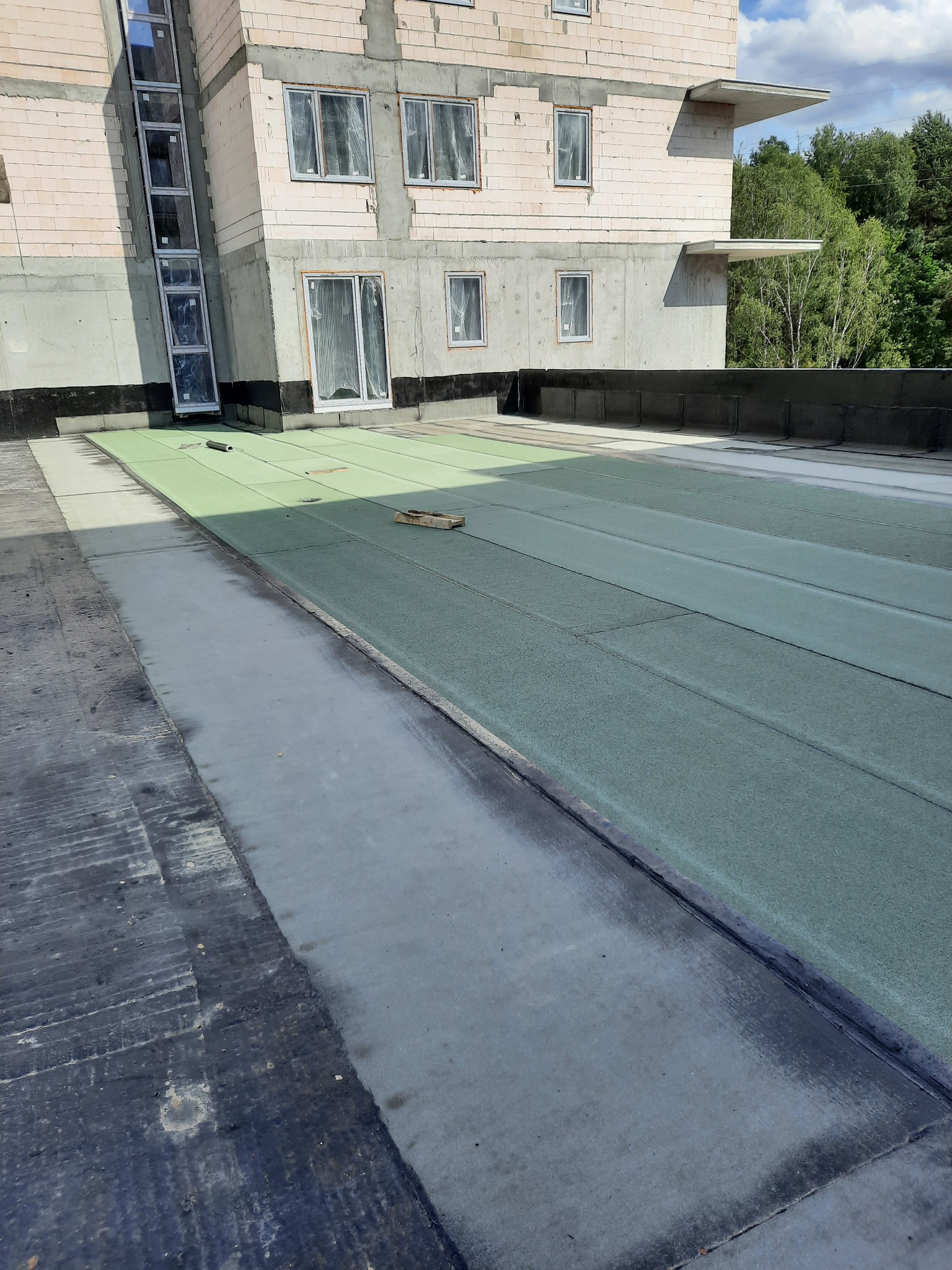 Niedokończone krycie dachu płaskiego z zielonej papy termozgrzewalnej na budynku w stanie surowym otwartym, widoczne drewniane kliny podtrzymujące papę.